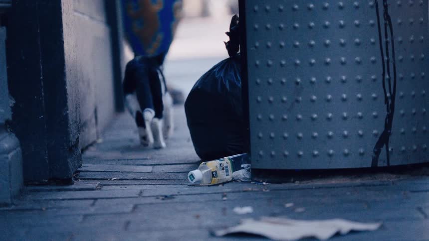A slow-motion shot from behind shows a black and white cat walking away down a littered city sidewalk