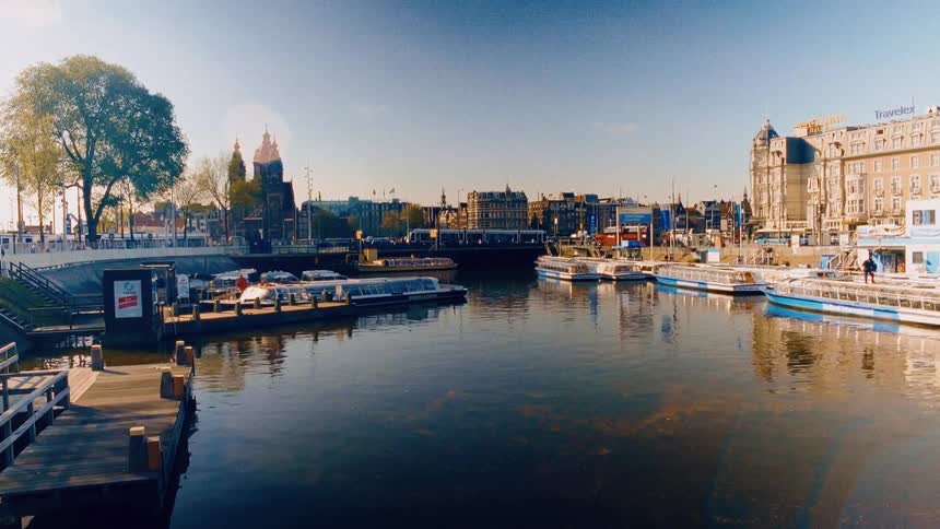 A sun-drenched, static view of a busy canal in Amsterdam, with tour boats moored at the docks and the historic city skyline in the background