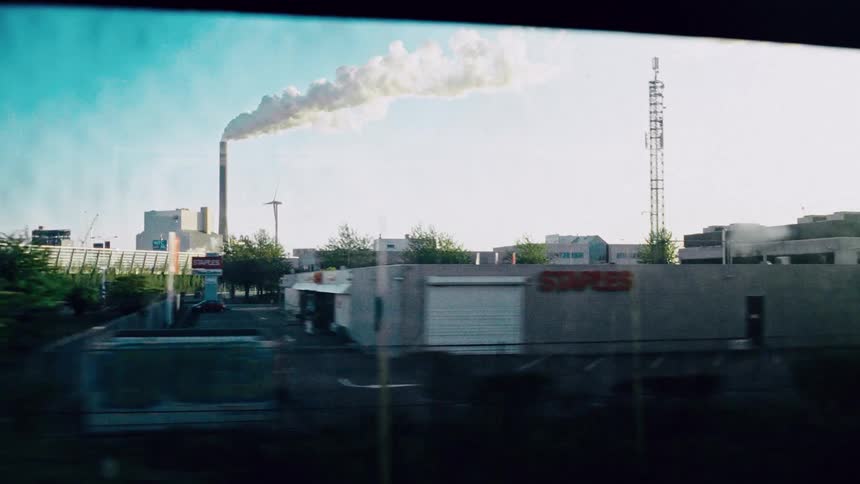 A view from a moving vehicle window captures an industrial plant with a tall smokestack emitting smoke against a bright sky