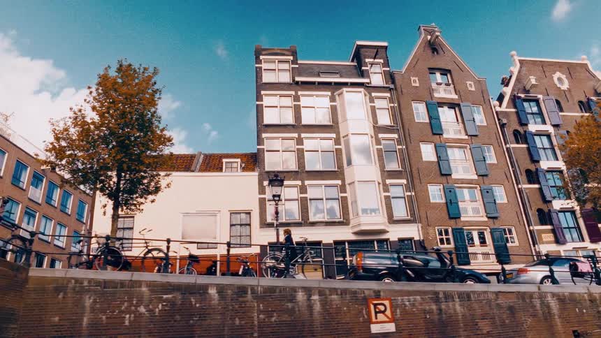 A cinematic, low-angle tracking shot from a boat captures the iconic architecture of Amsterdam's canal houses before passing under a bridge