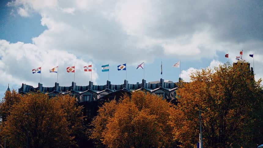 A slow pull-back shot reveals a grand historic building with numerous flags waving in the wind above bright orange autumn trees