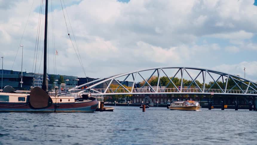 A tour boat passes under a modern arched pedestrian bridge over a river in a bustling city on a cloudy day