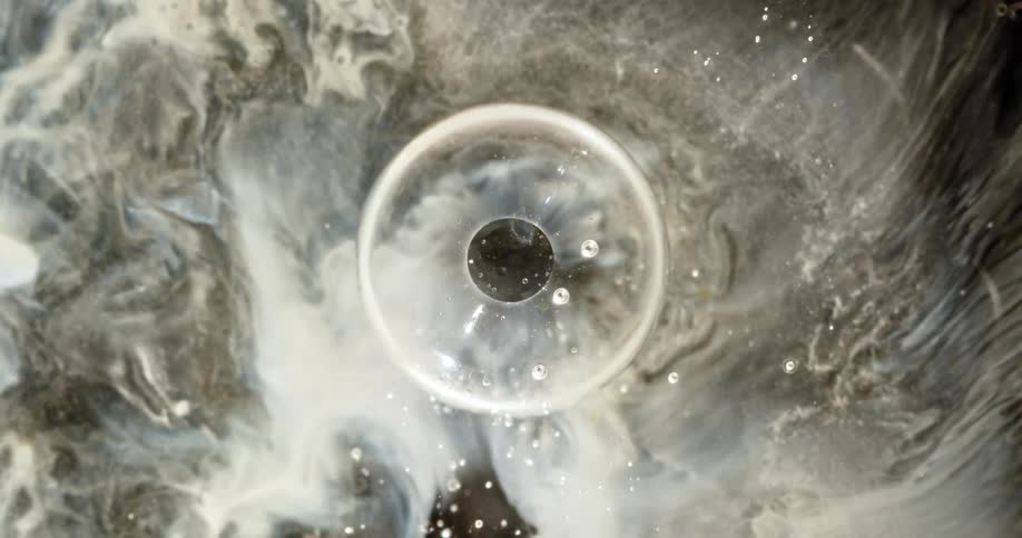 An abstract macro top-down shot shows a single bubble bursting in slow motion amidst a turbulent sea of milky white and black paint