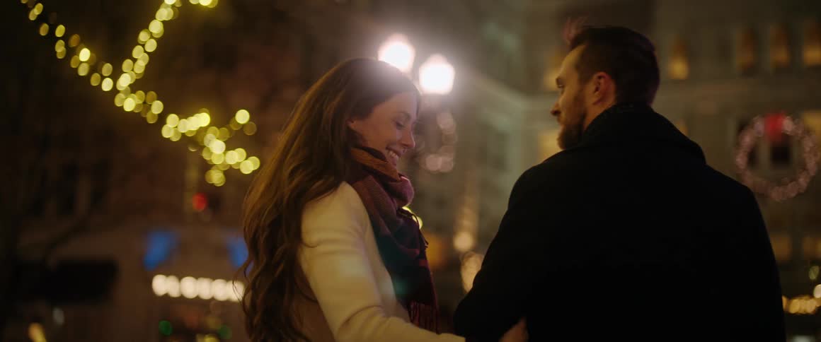A loving couple enjoys a romantic walk arm in arm down a city street beautifully decorated with festive lights at night