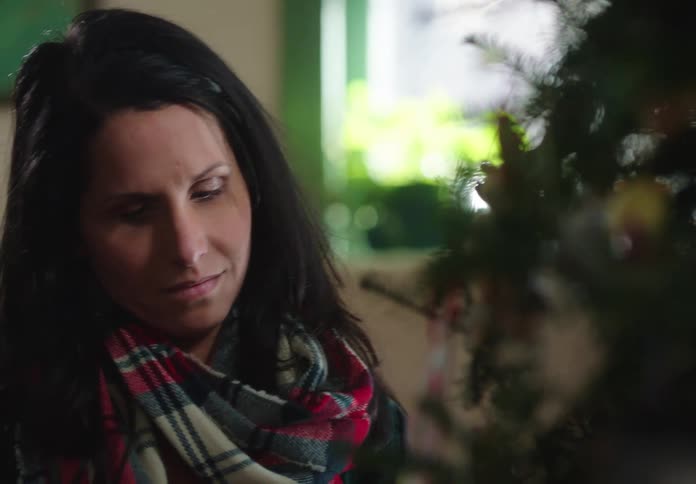 A woman with a plaid scarf smiles while carefully decorating a Christmas tree in her home