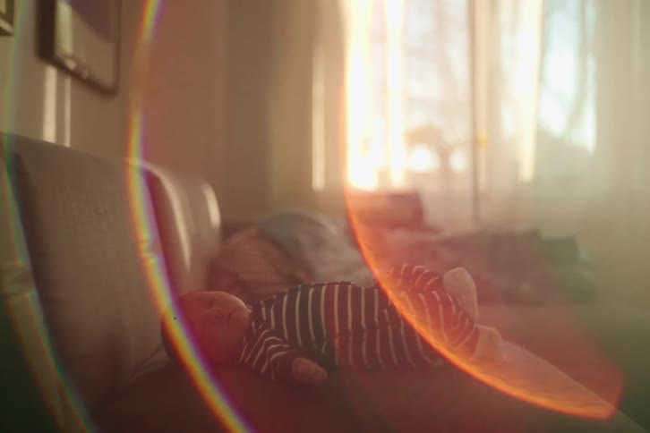 A newborn baby wearing a striped onesie rests peacefully on a couch in a sunlit room with dreamy lens flares