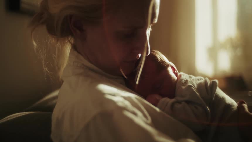 A tender moment as a new mother cuddles her sleeping newborn baby in the soft glow of sunlight