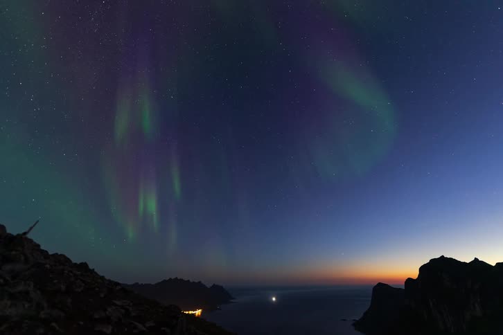 A breathtaking time-lapse shows the green and purple Northern Lights dancing across a star-filled sky above a tranquil fjord and mountain range at twilight
