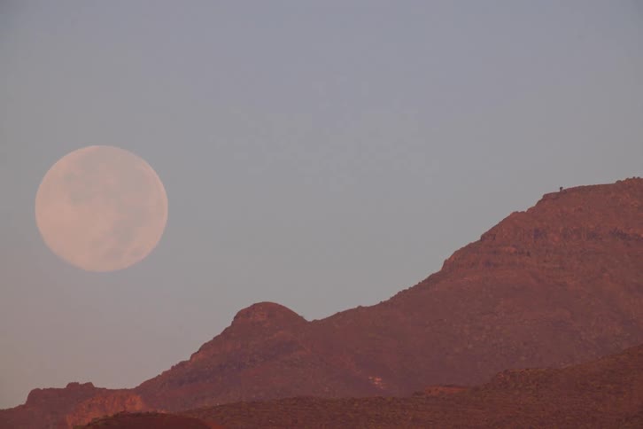 Time-lapse footage shows a large full moon setting behind a rocky mountain peak at dusk