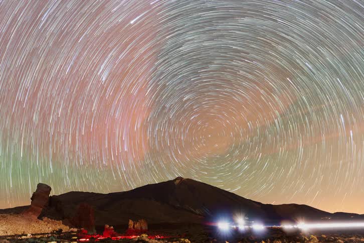 A stunning night-to-day time-lapse shows star trails spinning in concentric circles above a volcanic peak and unique rock formations