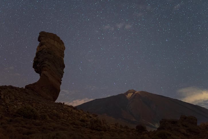 A majestic night timelapse captures stars and clouds moving across the sky above a prominent rock formation and a distant volcano