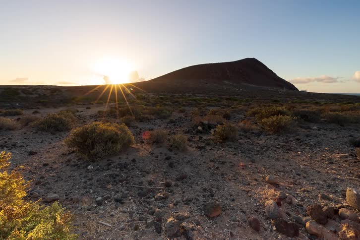 A beautiful timelapse shows the sun rising over a volcanic mountain in an arid, rocky landscape, casting long shadows across the terrain