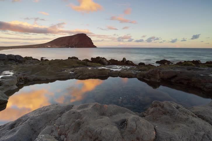 A scenic time-lapse shows a beautiful sunset over a rocky shoreline with a prominent red mountain, as the sky reflects in a calm tide pool