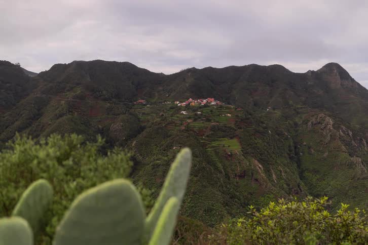 A rack focus shot reveals a picturesque village nestled in lush green mountains, starting from a close-up of a cactus