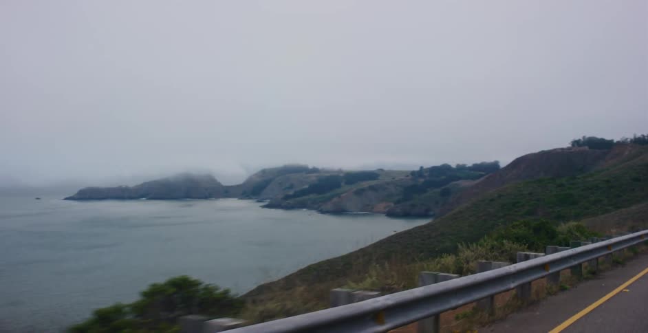 A car drives along a winding coastal highway, revealing a scenic view of the foggy ocean