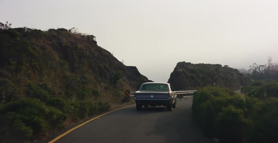 A vintage black muscle car drives down a winding road through the foggy coastal hills