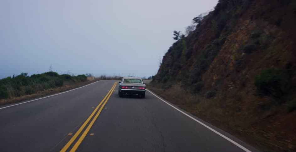 A vintage muscle car drives along a winding mountain road on an overcast day, followed from behind