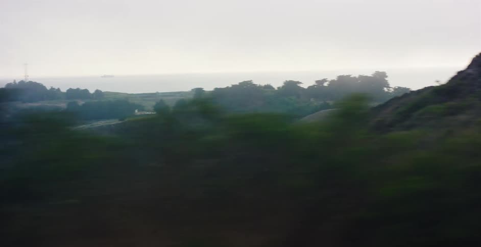 A dreamy, motion-blurred view from a moving vehicle captures the foggy coastline with rolling green hills