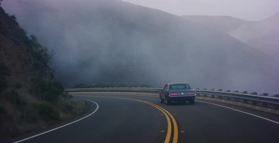A classic muscle car drives along a winding and foggy coastal highway at dusk