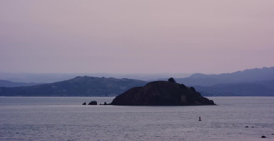 A solitary island rests in the calm bay waters under a hazy purple twilight sky with distant hills on the horizon