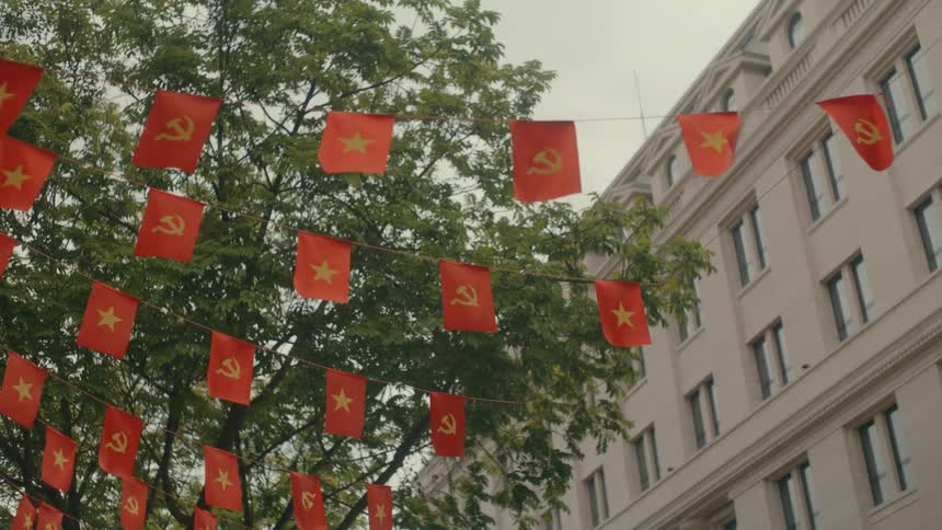 Red banners of the Vietnamese national flag and Communist Party flag wave in the wind against a backdrop of trees and a city building