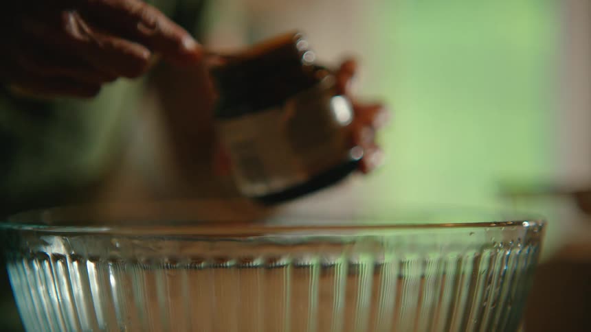 A baker carefully adds a spoonful of yeast to a steaming glass bowl in a cozy kitchen, captured in slow motion