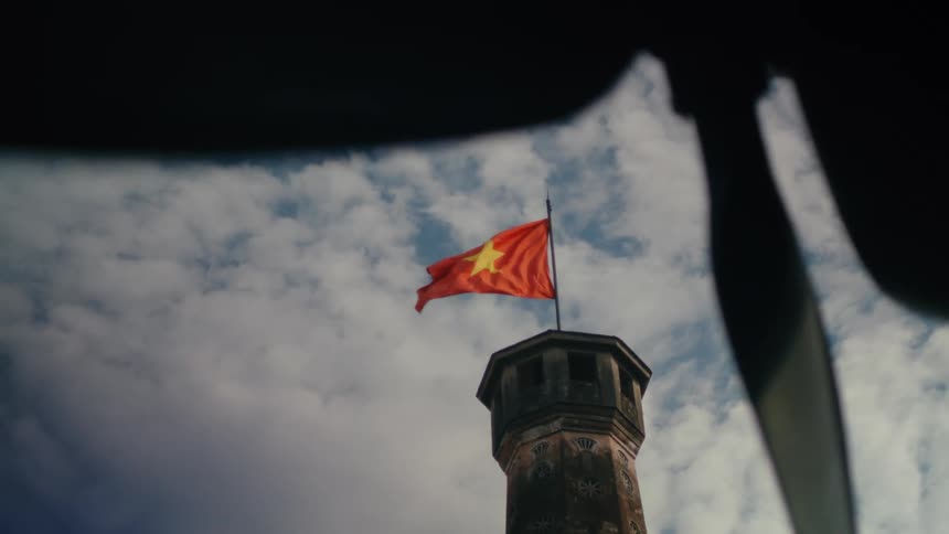 The Vietnamese flag waves proudly in the wind atop an ancient stone tower against a cloudy sky
