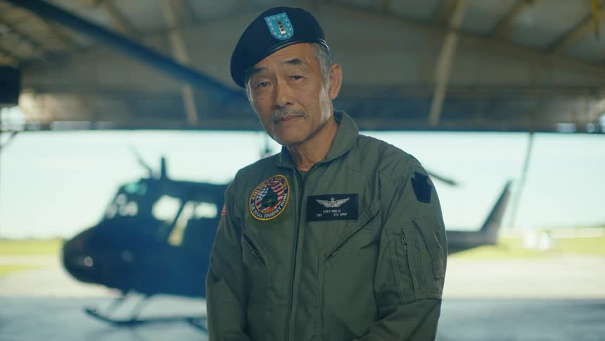 A stoic Asian American US Army veteran pilot stands proudly in a hangar with his helicopter in the background