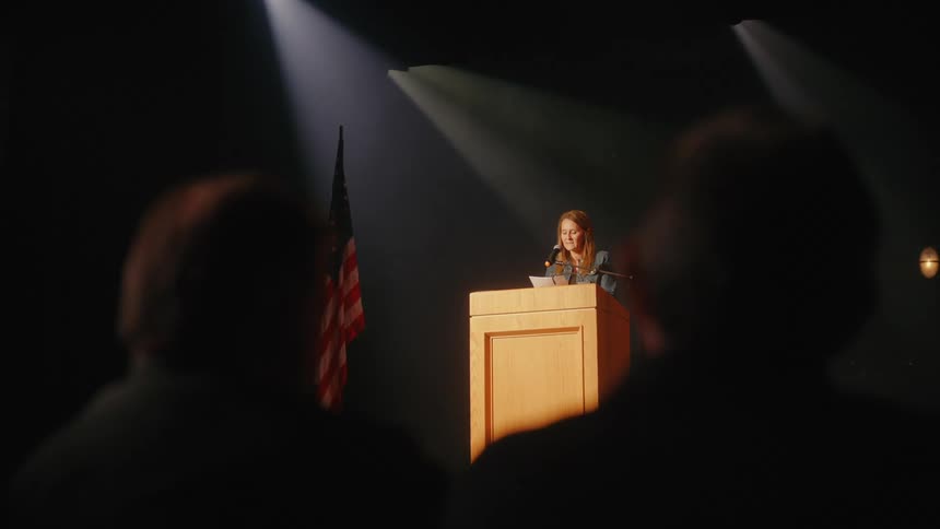 A female speaker delivers a formal speech from a podium on a dark stage, viewed from behind the audience