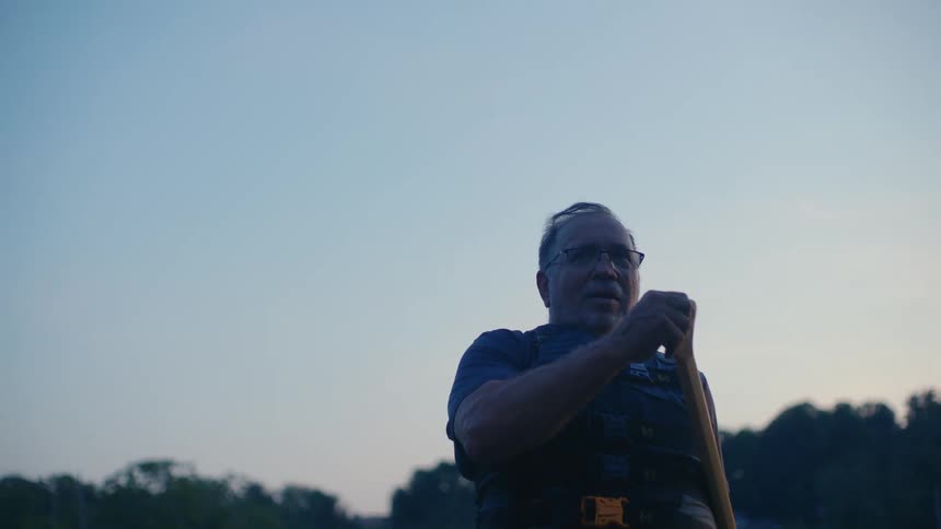 Low-angle shot of a serene older man in a life vest holding a paddle and reflecting on the water at dusk