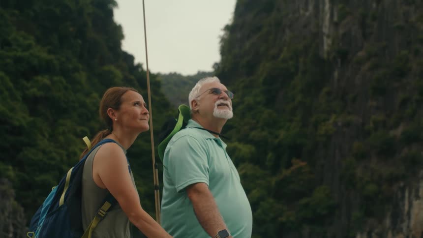 A mature couple with backpacks holds hands and looks up in awe at the majestic green mountains while on an adventure