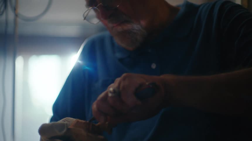 An elderly craftsman with glasses meticulously carves a small wooden sculpture in his dimly lit workshop