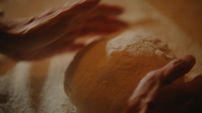 A close-up of elderly hands gently kneading a ball of bread dough in warm, soft light