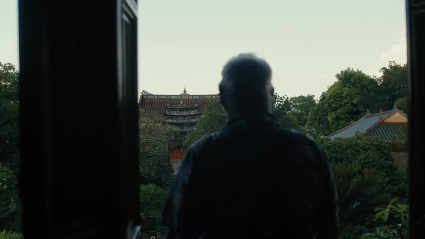 Silhouette of a pensive senior man looking out from a dark doorway over a tranquil Asian temple courtyard