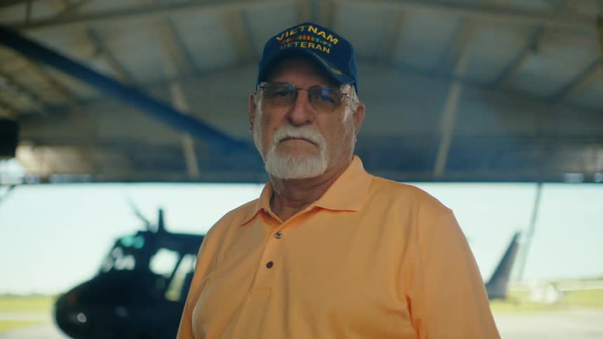 A portrait of a serious Vietnam War veteran standing in an aircraft hangar with a helicopter in the background