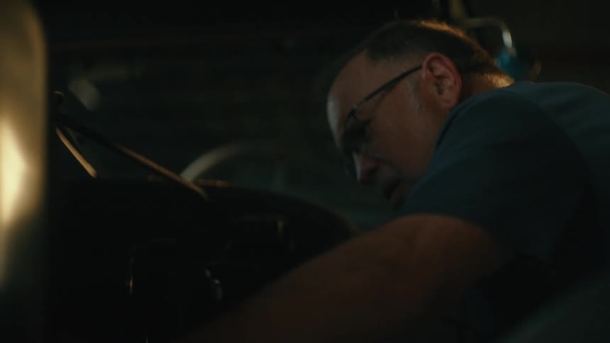 A focused mechanic with glasses works diligently on the engine of a vehicle inside a dimly lit garage