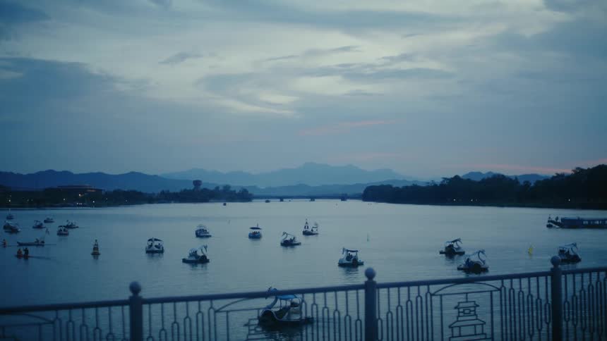 A serene blue hour view from a bridge overlooking paddle boats floating on a wide river with mountains in the distant background