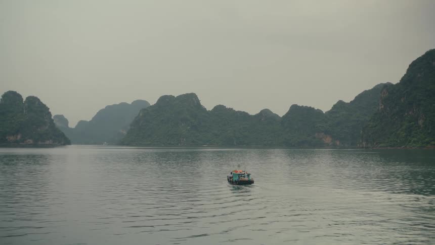 A small local boat travels through the calm waters of a vast bay surrounded by large, green, karst islands on a misty, overcast day