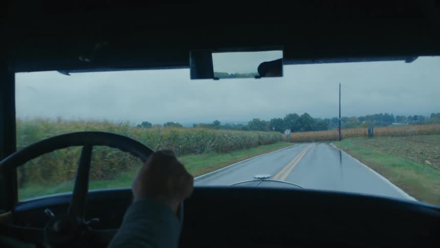 Point of view from inside a vintage car driving down a wet country road past cornfields on an overcast day