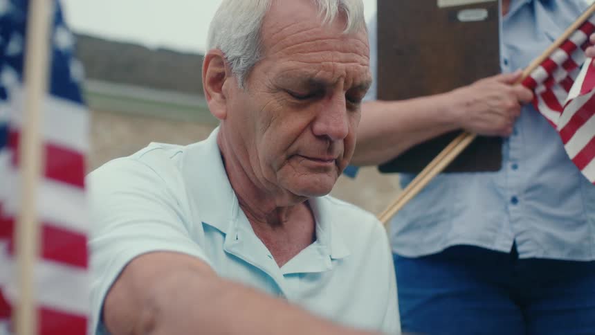 A solemn elderly man reflects quietly while honoring a memorial, surrounded by patriotic American flags