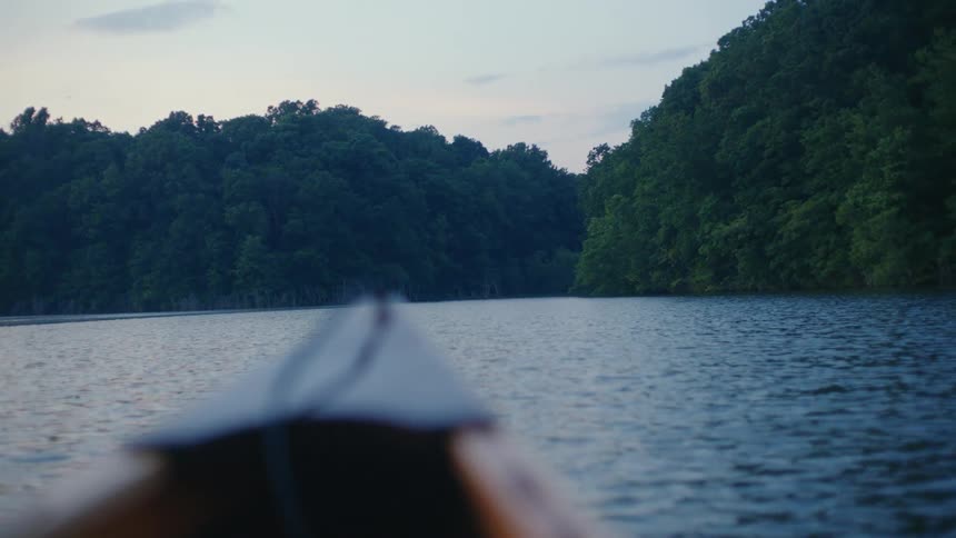 A peaceful point-of-view from the bow of a canoe floating on a calm lake as the focus shifts to the lush forest shoreline at twilight