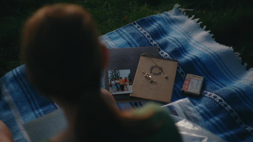A person sits on a blanket in the grass, surrounded by cherished keepsakes and old photographs, reminiscing about past memories