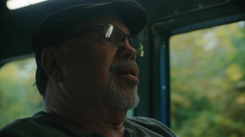 A close up on the pensive face of a senior man in a flat cap and glasses as he rides a vehicle through the countryside