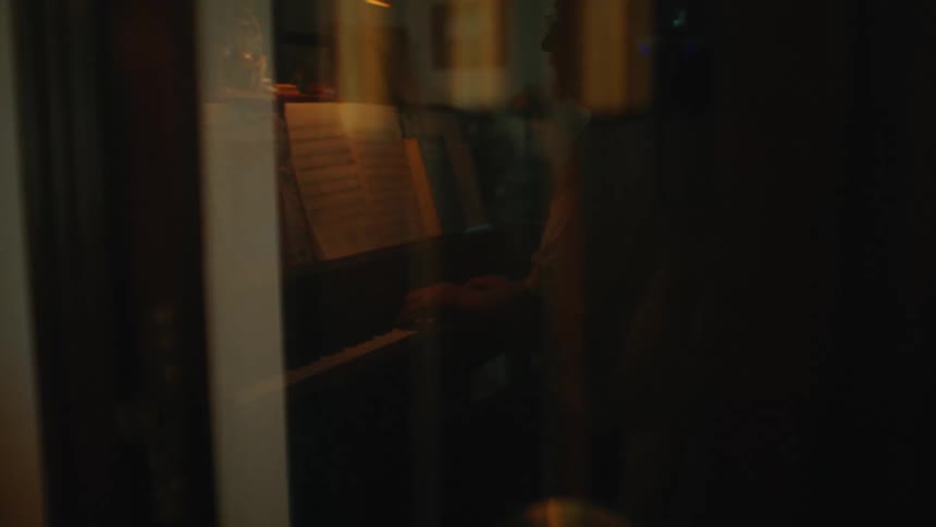 A moody reflection of a musician playing the piano is seen in the glass of a grandfather clock as the pendulum swings