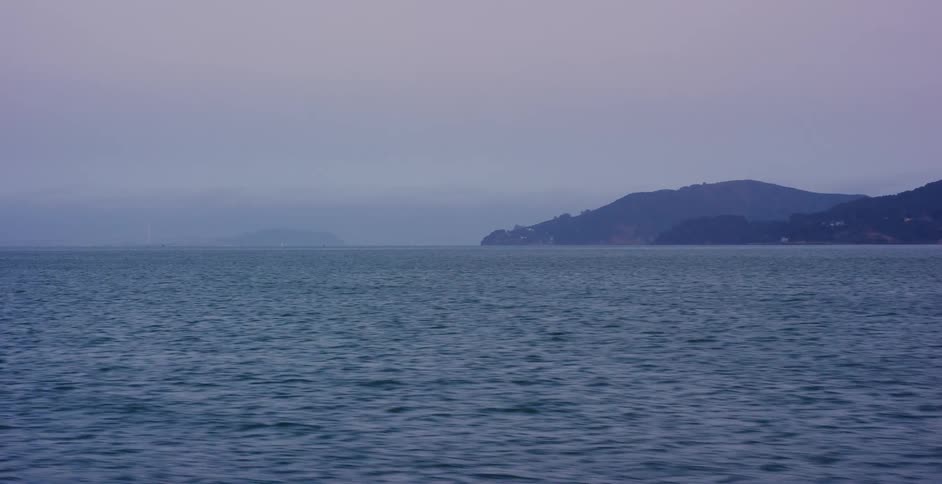 A static shot of a calm bay with misty hills in the background under an overcast sky