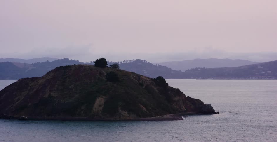 A gloomy point-of-view shot from a moving vehicle on a bridge shows a small island in a large bay on an overcast day
