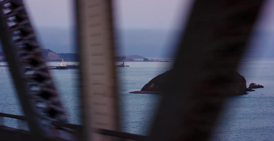 Driving over a bridge provides a parallax view of a small island and an industrial shipping port in the bay on an overcast day