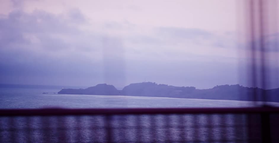 A dreamy, purple-hued tracking shot of a foggy coastline seen from a vehicle crossing a large bridge