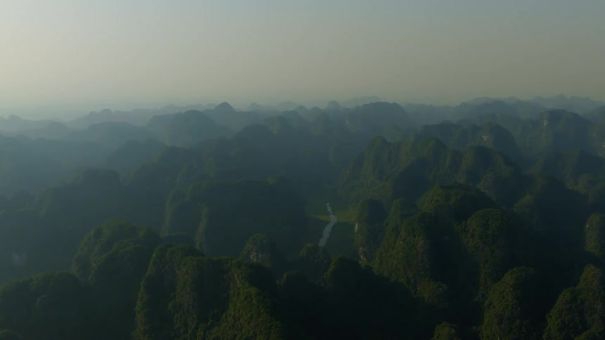 An expansive aerial view of a hazy, green mountain range with a winding river valley below at sunrise