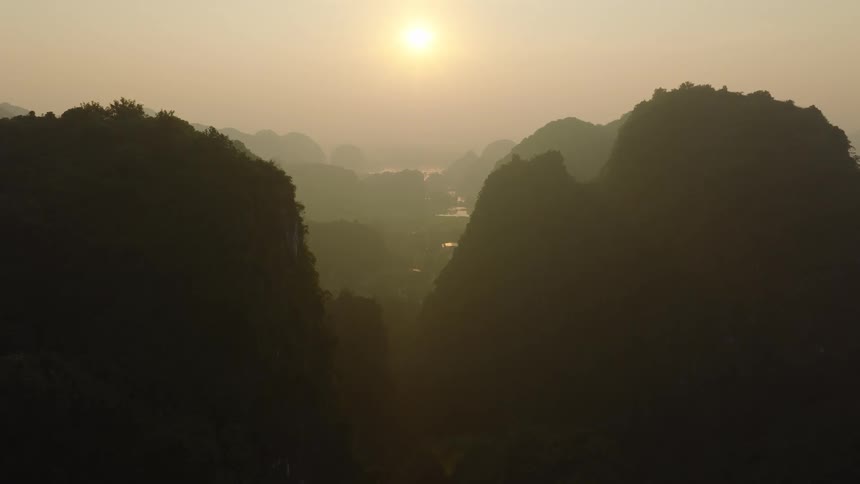A static aerial view shows the sun rising over a vast, hazy mountain range, silhouetting the peaks against the golden sky